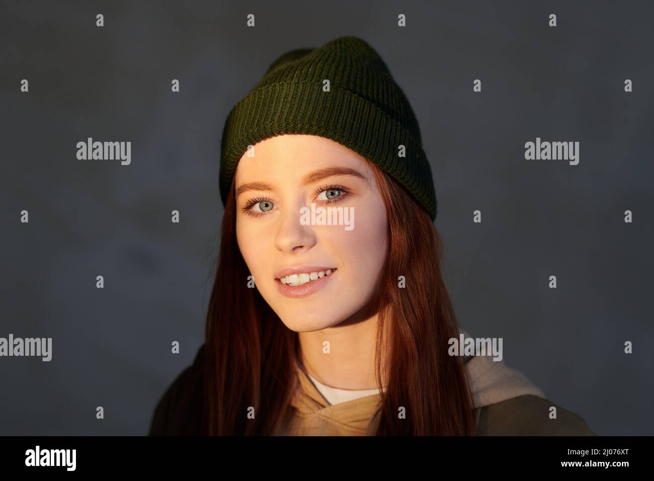 Horizonatal close-up shot of attractive young woman with darl red hair wearing cap smiling at camera, gray background Stock Photo