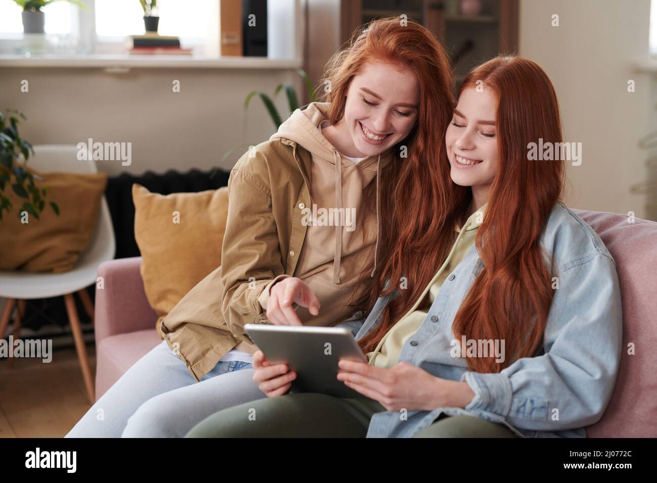 Portrait of red-haired twin sisters spending free time together sitting on sofa in living room enjoying surfing Internet using digital tablet Stock Photo