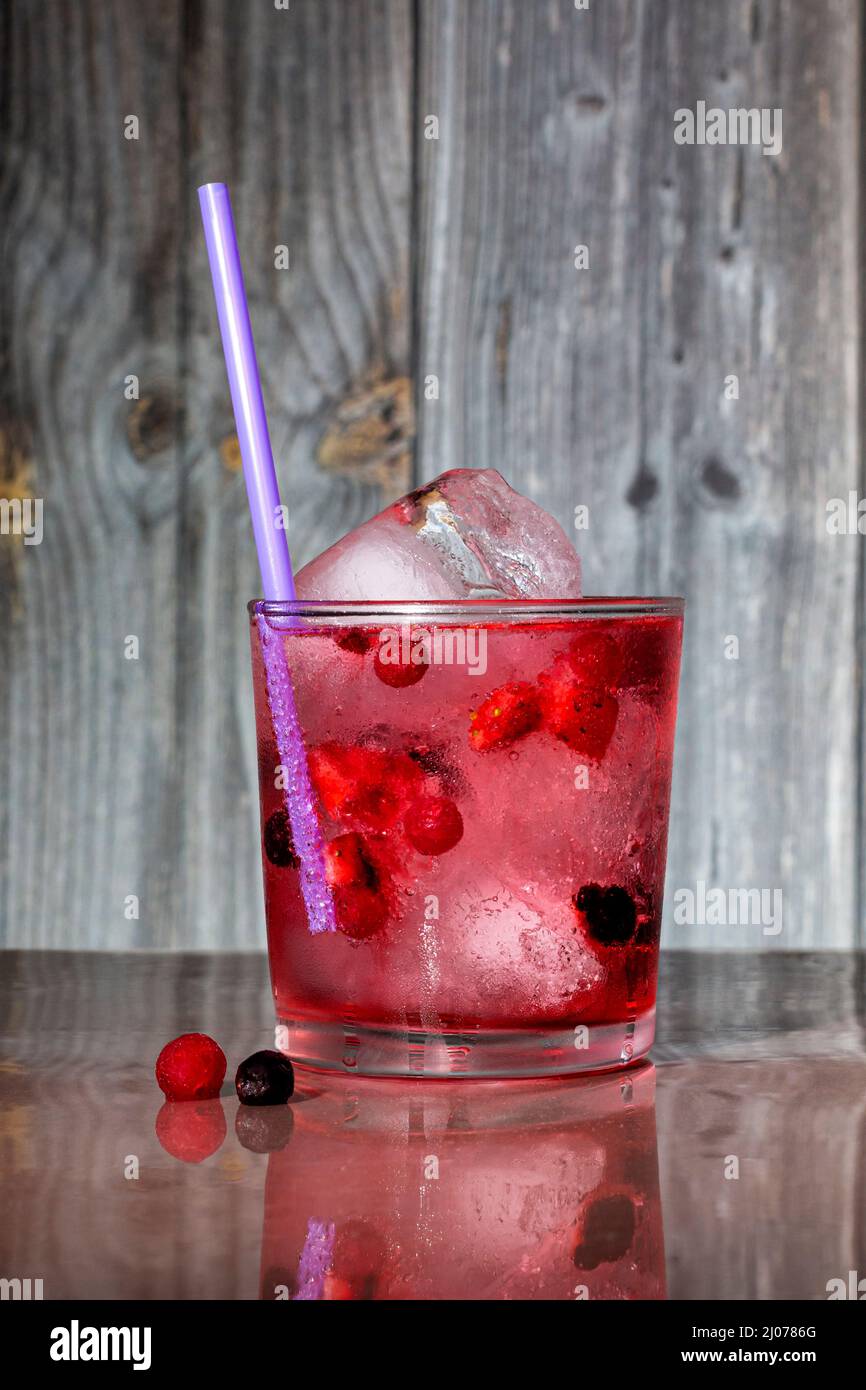 A cylindrical glass with a combination of gin, tonic, ice and red fruits with a straw. It is placed on a brown table where it reflects and in the back Stock Photo