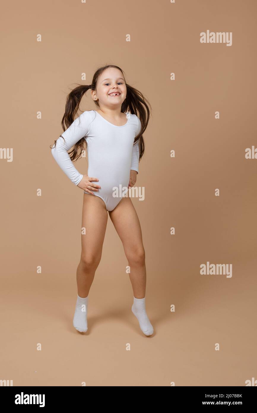 Portrait of young adorable happy smiling girl with long dark hair in white training swimsuit and socks jumping on brown background, posing, having fun Stock Photo