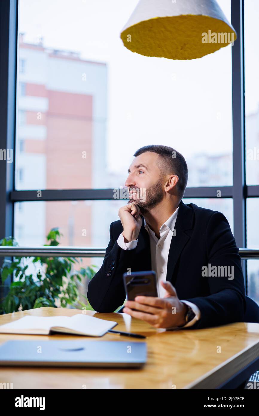 Confident male businessman in a business suit sits at a table with a phone and works remotely Stock Photo