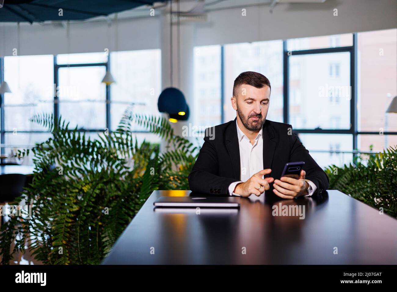 Confident male businessman in a business suit sits at a table with a phone and works remotely Stock Photo
