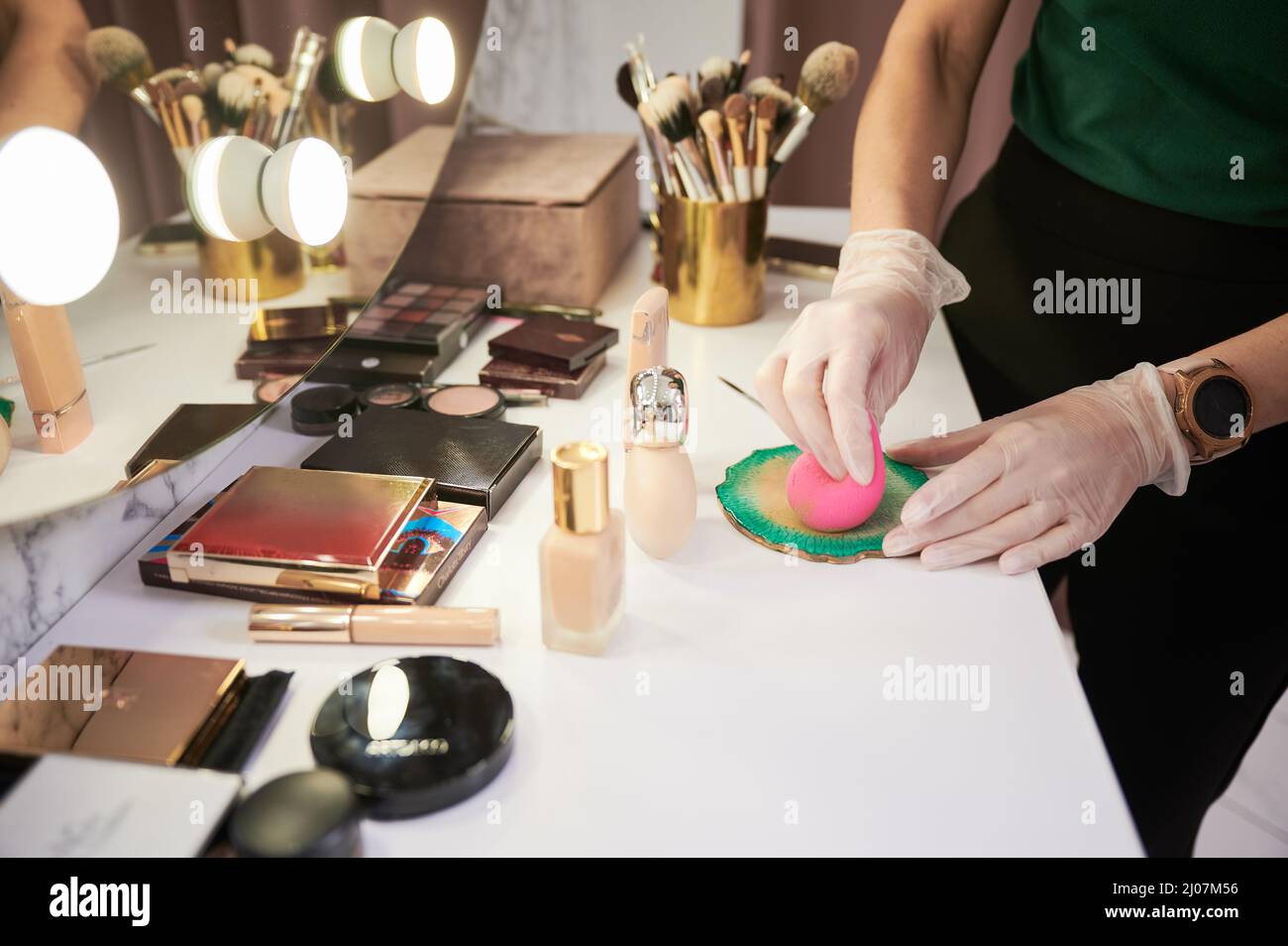 Close up of woman hands in sterile gloves preparing blending beauty sponge for applying foundation. Female makeup artist standing by dressing table with various cosmetics in beauty salon. Stock Photo