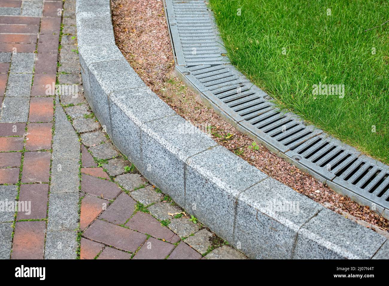 iron drainage grate on the walkway side with green turf lawn and stone pebbles at the granite curb along the pedestrian pavement of stone brick tiles, Stock Photo