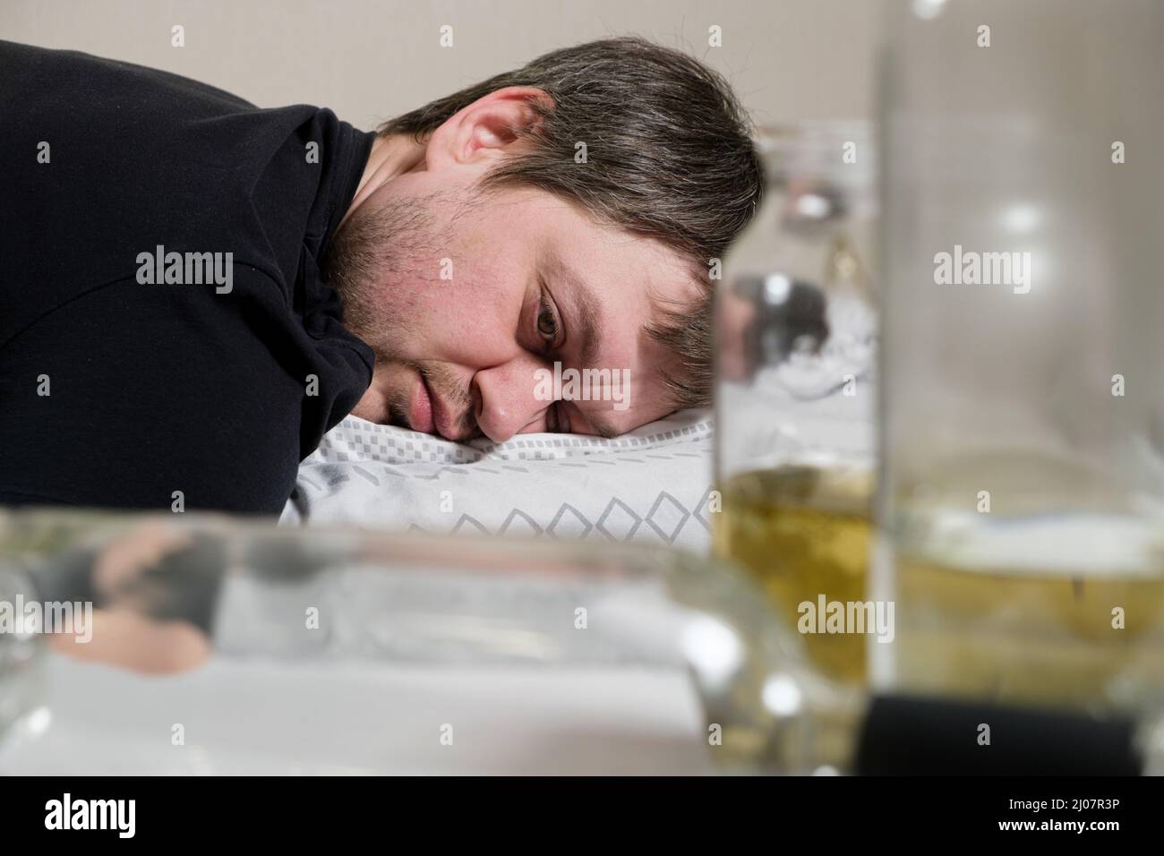 A drunken alcoholic man lies on the bed, empty bottles on the table. Alcoholism, alcohol dependence as a social problem Stock Photo