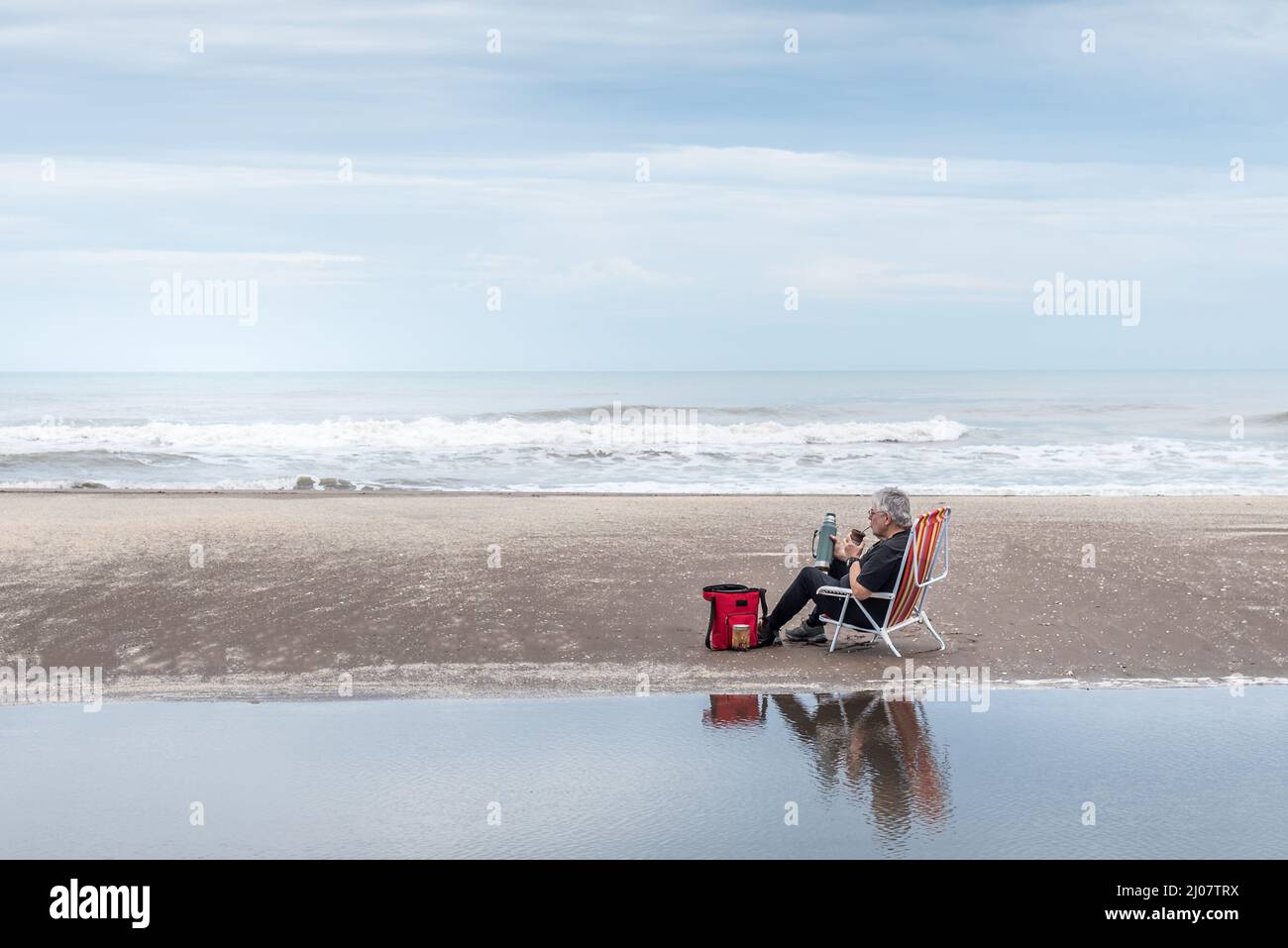 Mature man with gray hair and glasses sitting on a beach chair drinking mate all reflected in the water and in the background the waves of the sea. Stock Photo