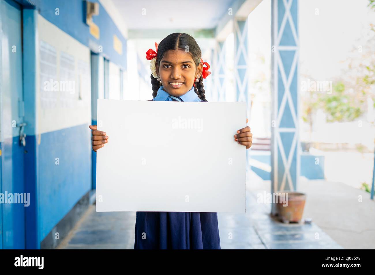 Happy girl kid in uniform showing white empty board or placard by looking at camera at school corridor - concept of education, announcement and Stock Photo
