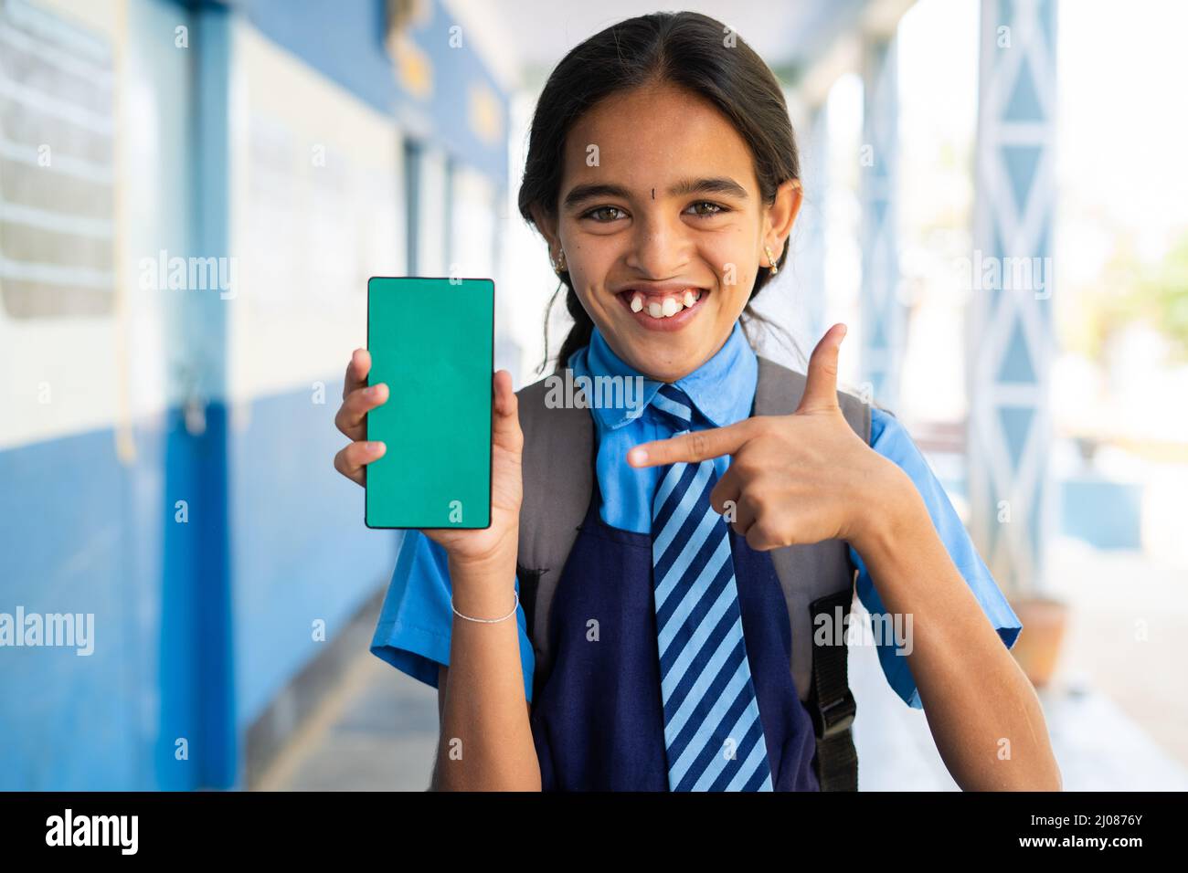 Smiling school girl kid in uniform showing green screen mobile phone by pointing finer while looking camera at corridor - concept of education Stock Photo