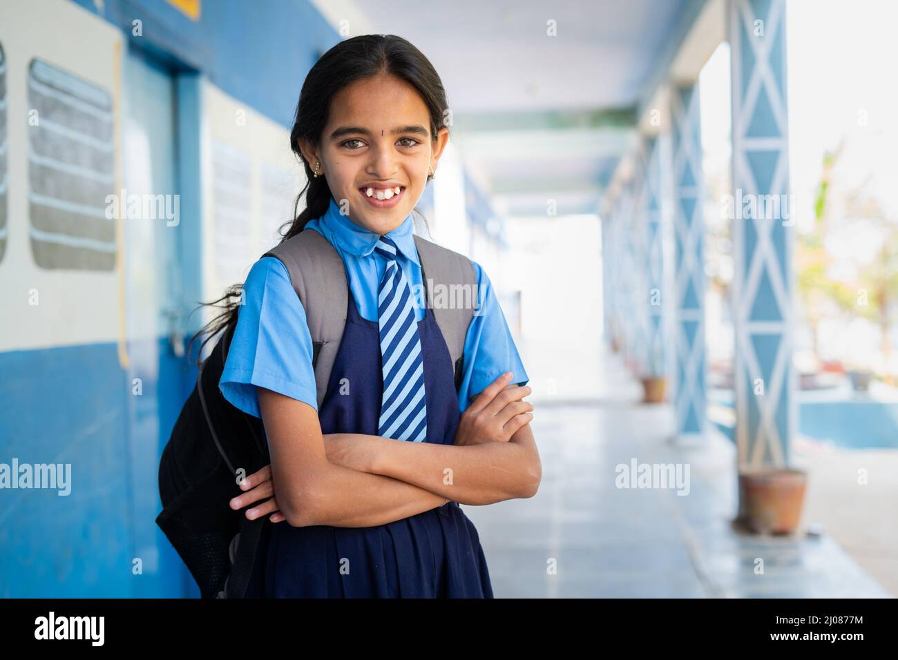 Happy smiling girl kid in school uniform confidently standing at corridor with arms crossed by looking at camera - concept of education, knowledge and Stock Photo