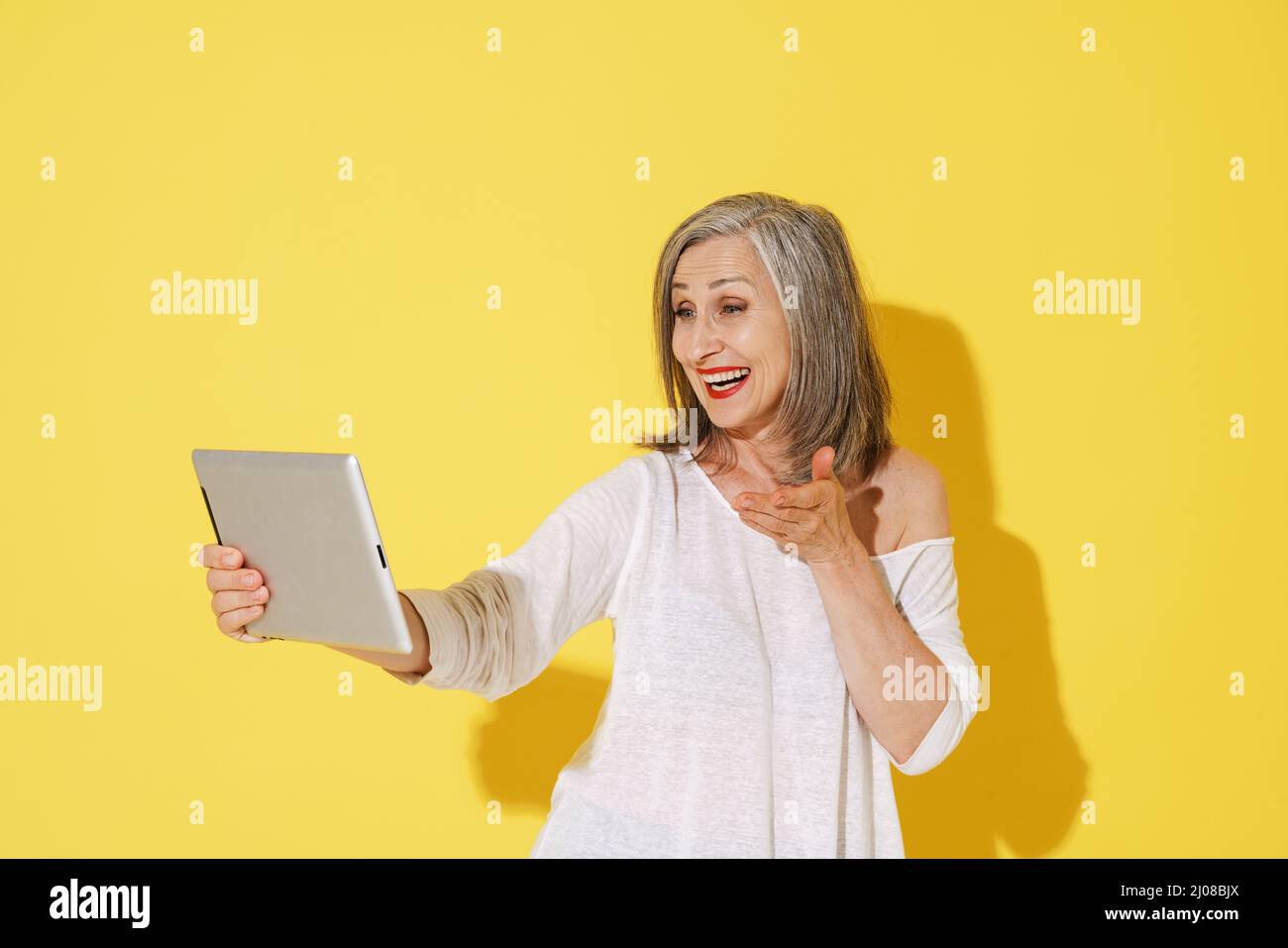 White senior woman gesturing while using tablet computer isolated over yellow wall Stock Photo