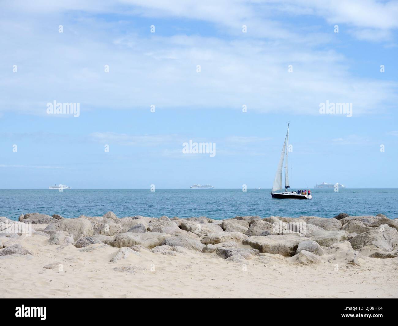 A sailing yacht passing close to the shore with three anchored cruise liners in the distance Stock Photo