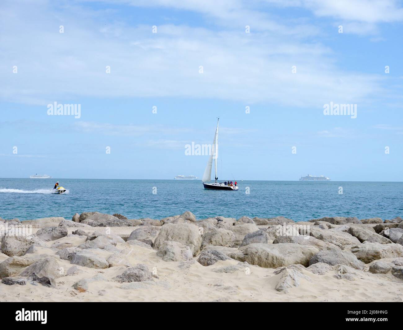 A sailing yacht passing close to the shore with three anchored cruise liners in the distance and a jet ski passing by Stock Photo