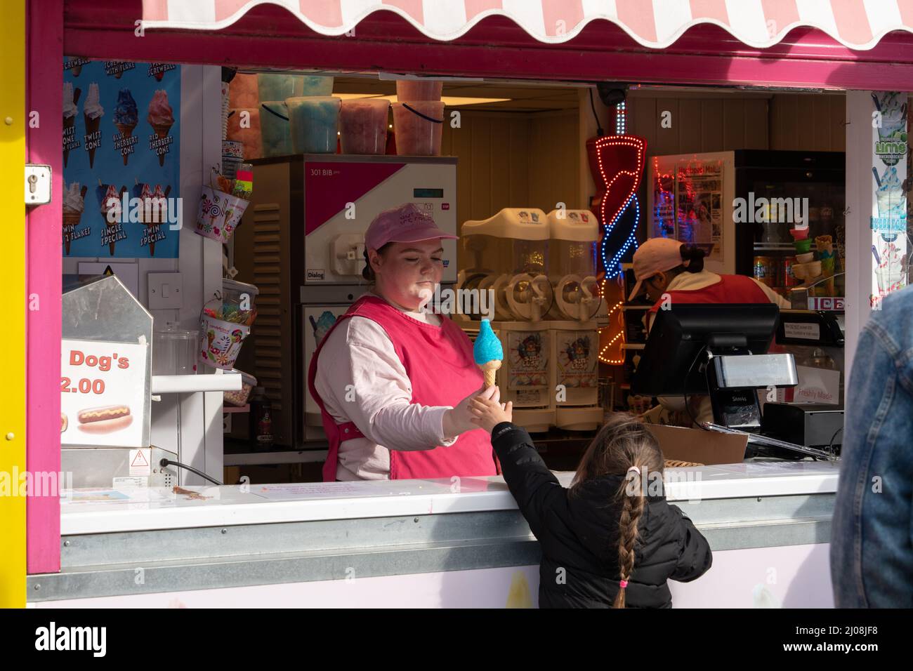 An ice cream seller at a seaside kiosk passes a blue ice cream in a cone to a young girl with a long plait. Staycations continue to be popular in UK. Stock Photo