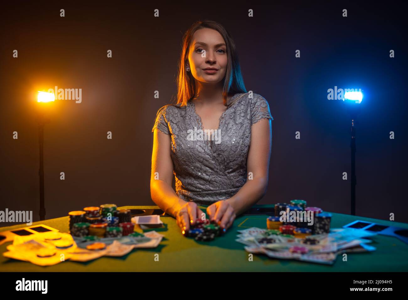 Unemotional woman siting at the table in casino and play poker. Lucky player on smoky background with lamps. Concept of gambling. Stock Photo