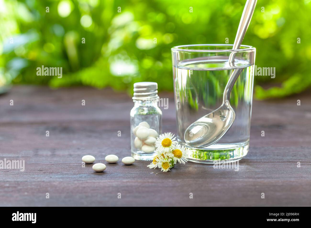 Homeopathic concept. Drinking glass with water, dragees and chamomile on a wooden table. Bright green in the background. Focus on the foreground. Stock Photo