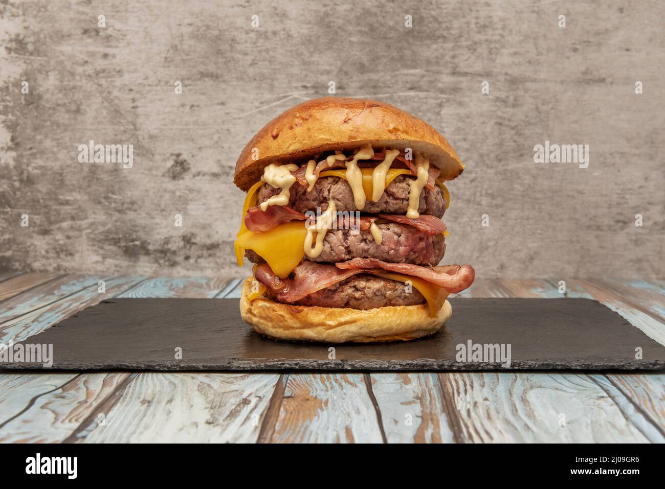 a beastly three meat burger with lots of fried bacon, cheddar cheese and sauce to give you a heart attack and a good bite on a black slate board and b Stock Photo