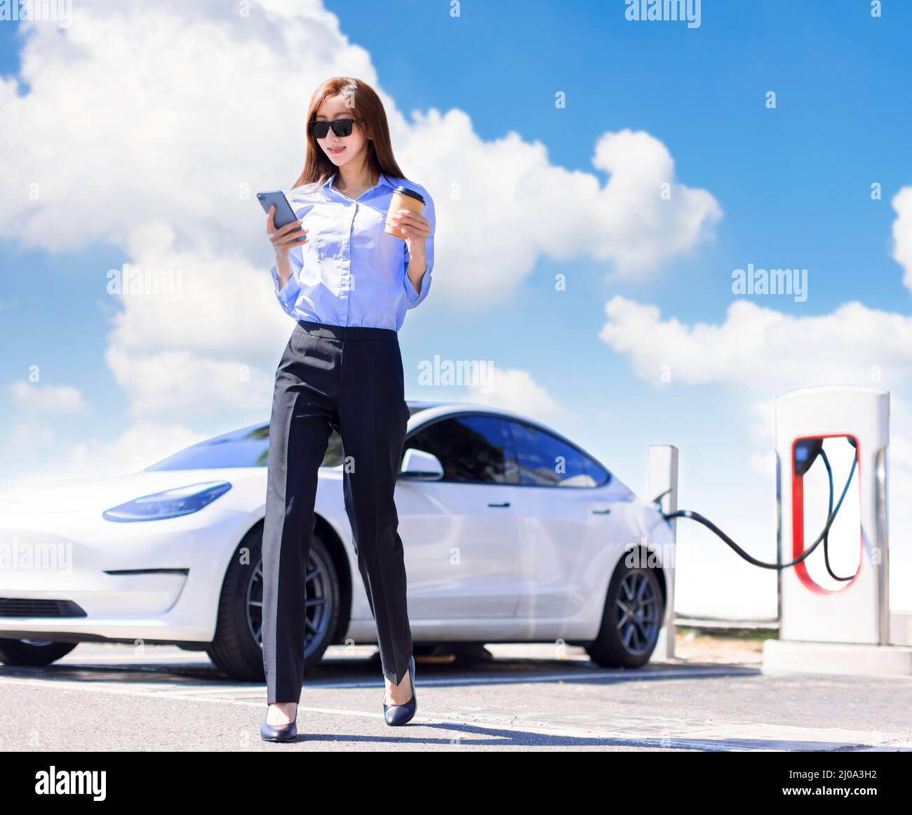 Smiling  business woman walking on city parking near electric car, charging automobile battery from small city station, drinking coffee and watching t Stock Photo