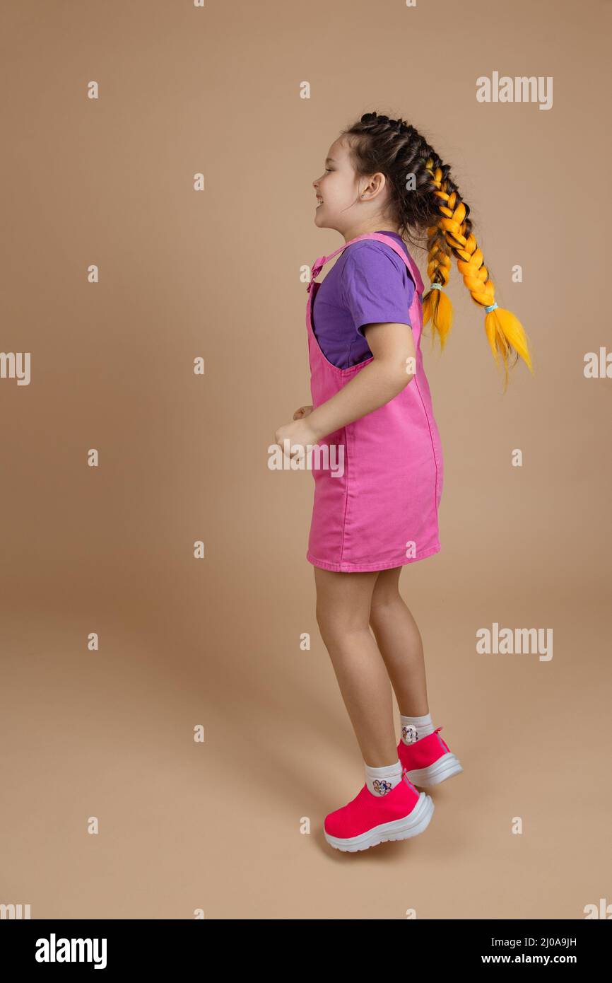 Enthusiastic blissful female child with two kanekalon pigtails of yellow color having fun jumping dressed in pink jumpsuit, purple t-shirt and pink Stock Photo