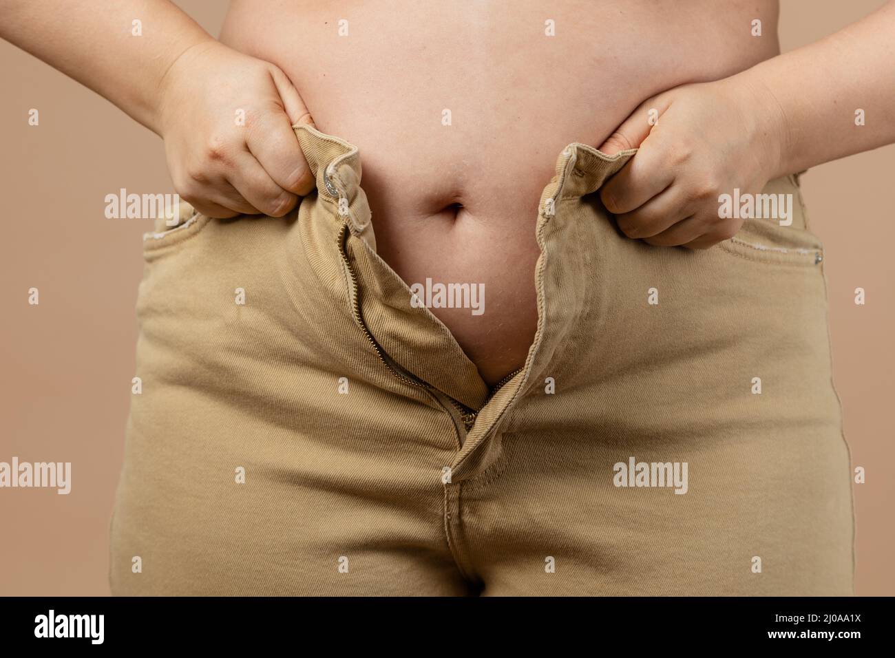 Female flabby big tummy preventing her from putting jeans on on beige background. Visceral fat. Body positive. Sudden weight gain. Tight little Stock Photo