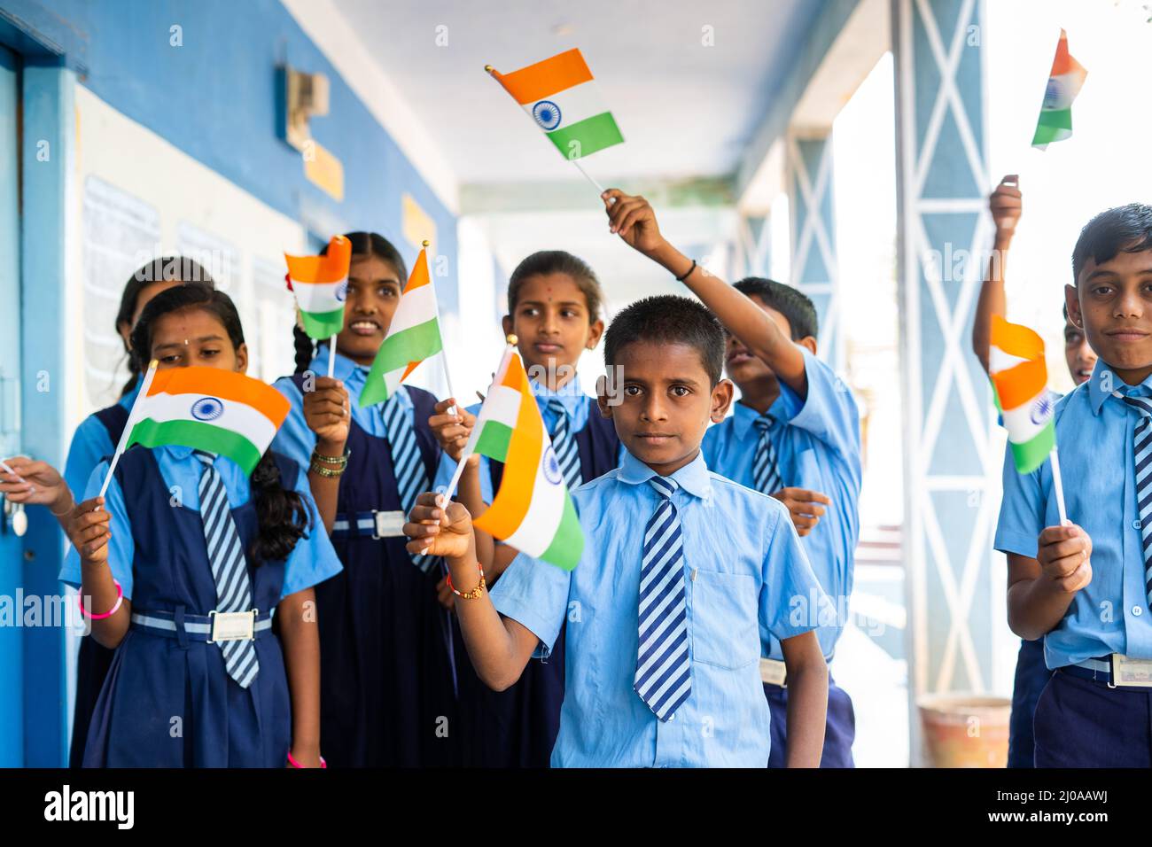 Happy kids in uniform waving indian flag by looking camera at school corridor - concept of independence or republic day celebration, patriotism and Stock Photo
