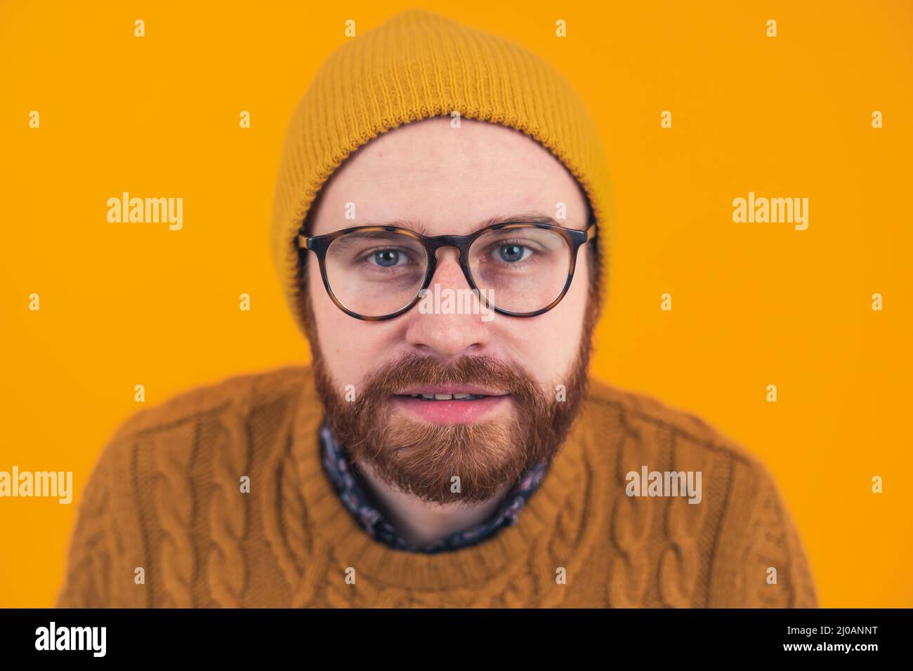 Headshot attractive young bearded Caucasian man in glasses and hat looking with interest at the camera hipster isolated selective focus on the head orange background studio shot . High quality photo Stock Photo