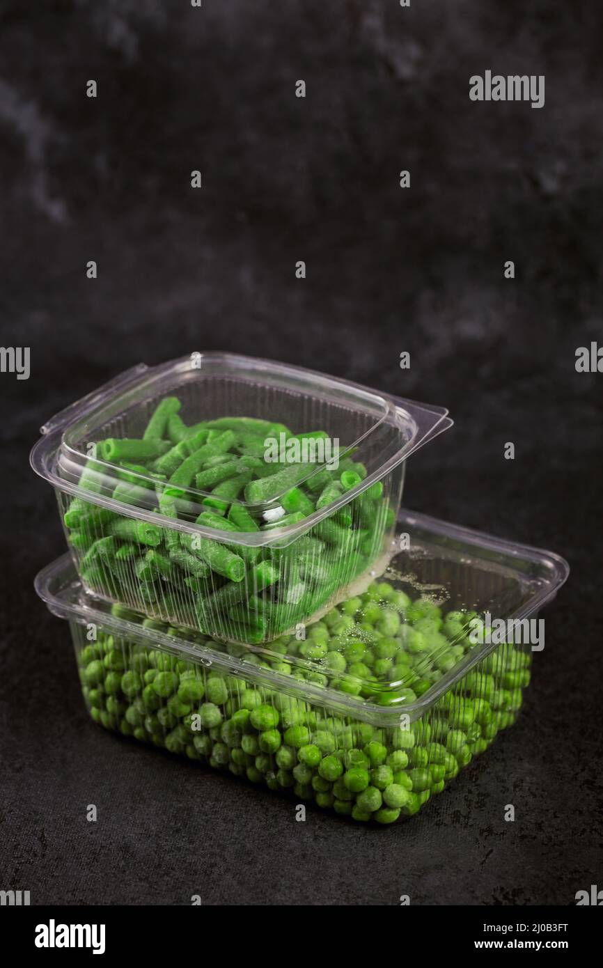 Plastic container with different organic deep frozen vegetables on black table. Green peas and