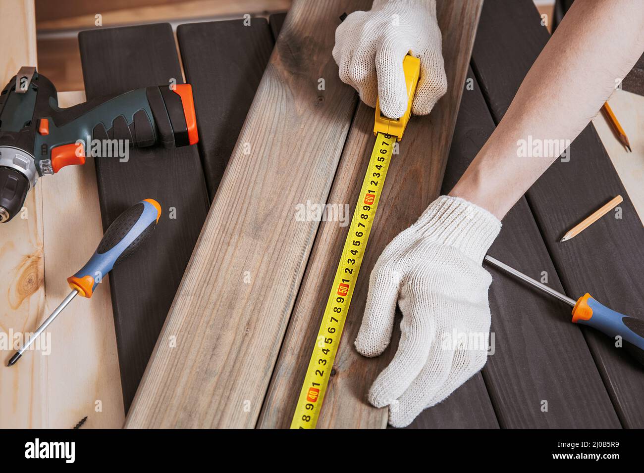 Hands in gloves of joiner in carpentry. Carpenter is measuring length of wood planks or timbers by measuring tape or ruler. Carpenter workspace, craft Stock Photo