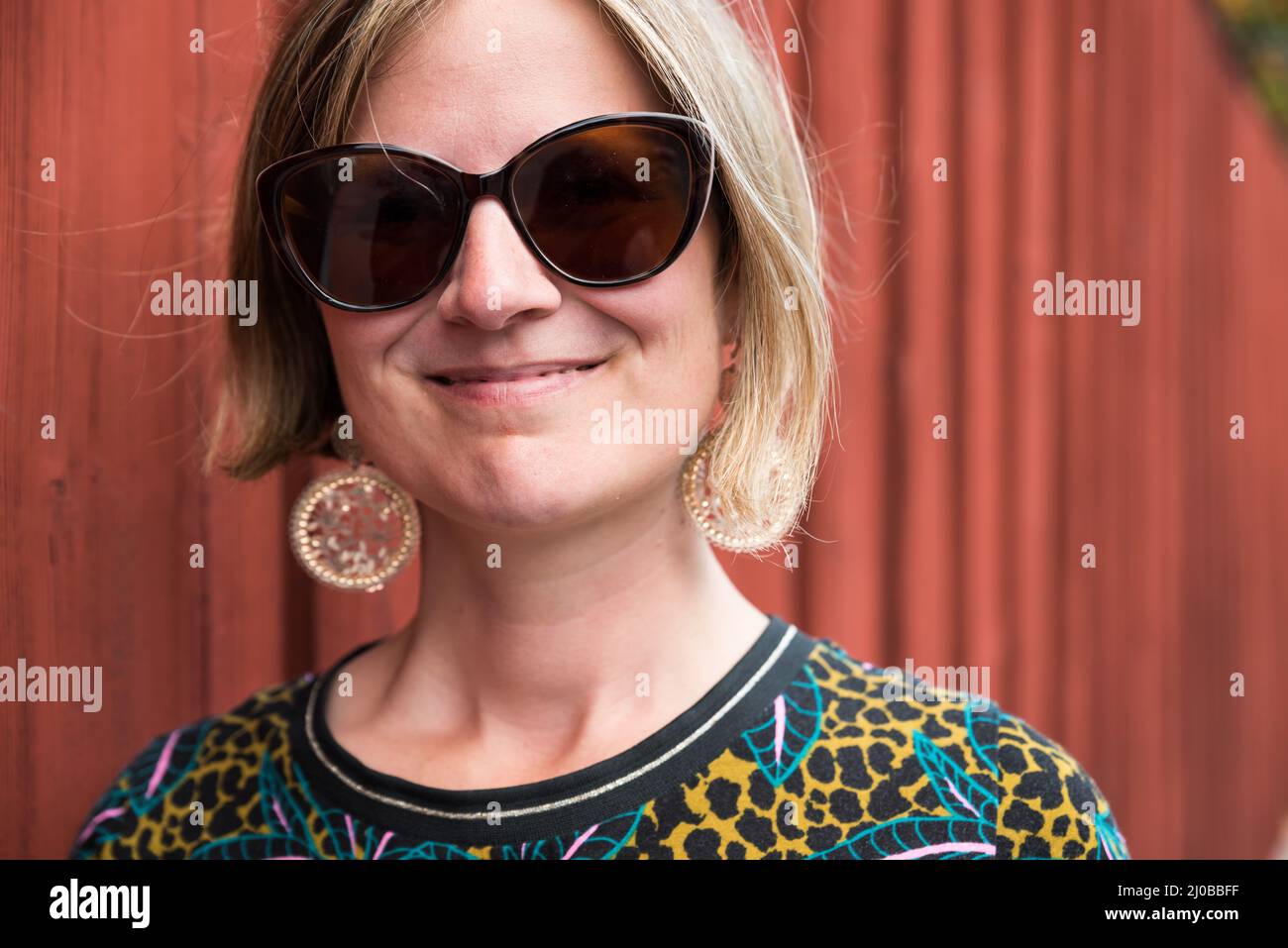 Portrait of an attractive thrity year old woman with sunglasses standing against a typical Falun red wall, Falun, Dalarna, Sweden Stock Photo