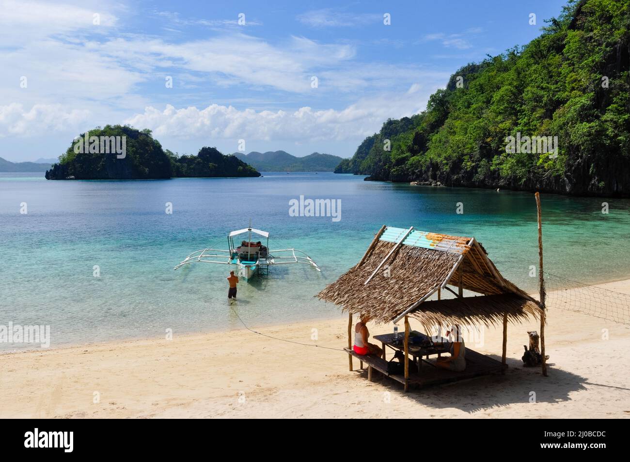 Bald Man pulling wooden traditional filipino boat while two women rest in bamboo hut at white sand beach Stock Photo