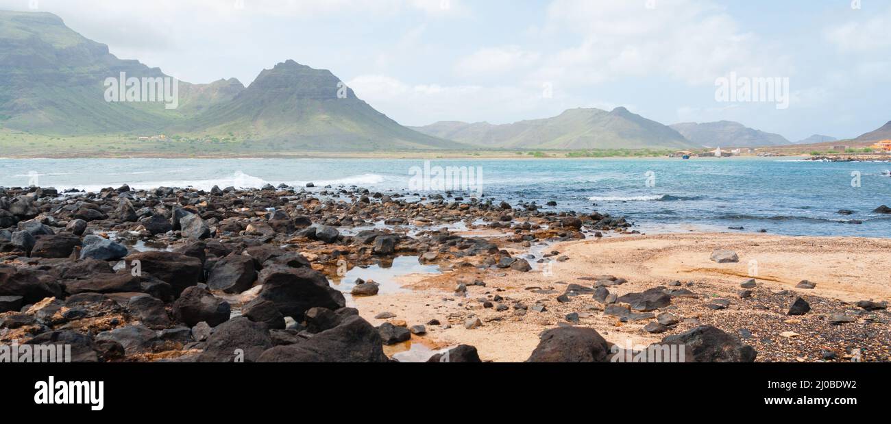 Black rock stone sand beach coast in front of blue sea with mountain background Stock Photo