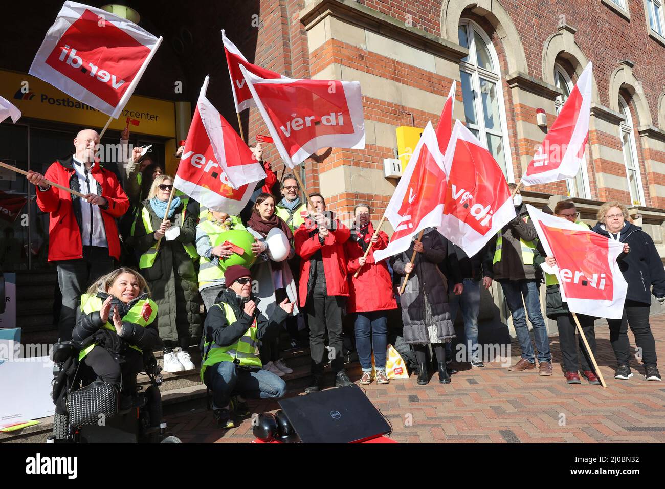 Hamburg, Germany. 18th Mar, 2022. Strikers stand together with flags at a rally in front of a closed Postbank branch. The trade union Verdi calls the 15,000 employees of Postbank to a nationwide strike starting today. Called to the walkouts from Friday, March 18, 2022, are employees in all Postbank branches, call centers and Postbank locations. Credit: Bodo Marks/dpa/Alamy Live News Stock Photo