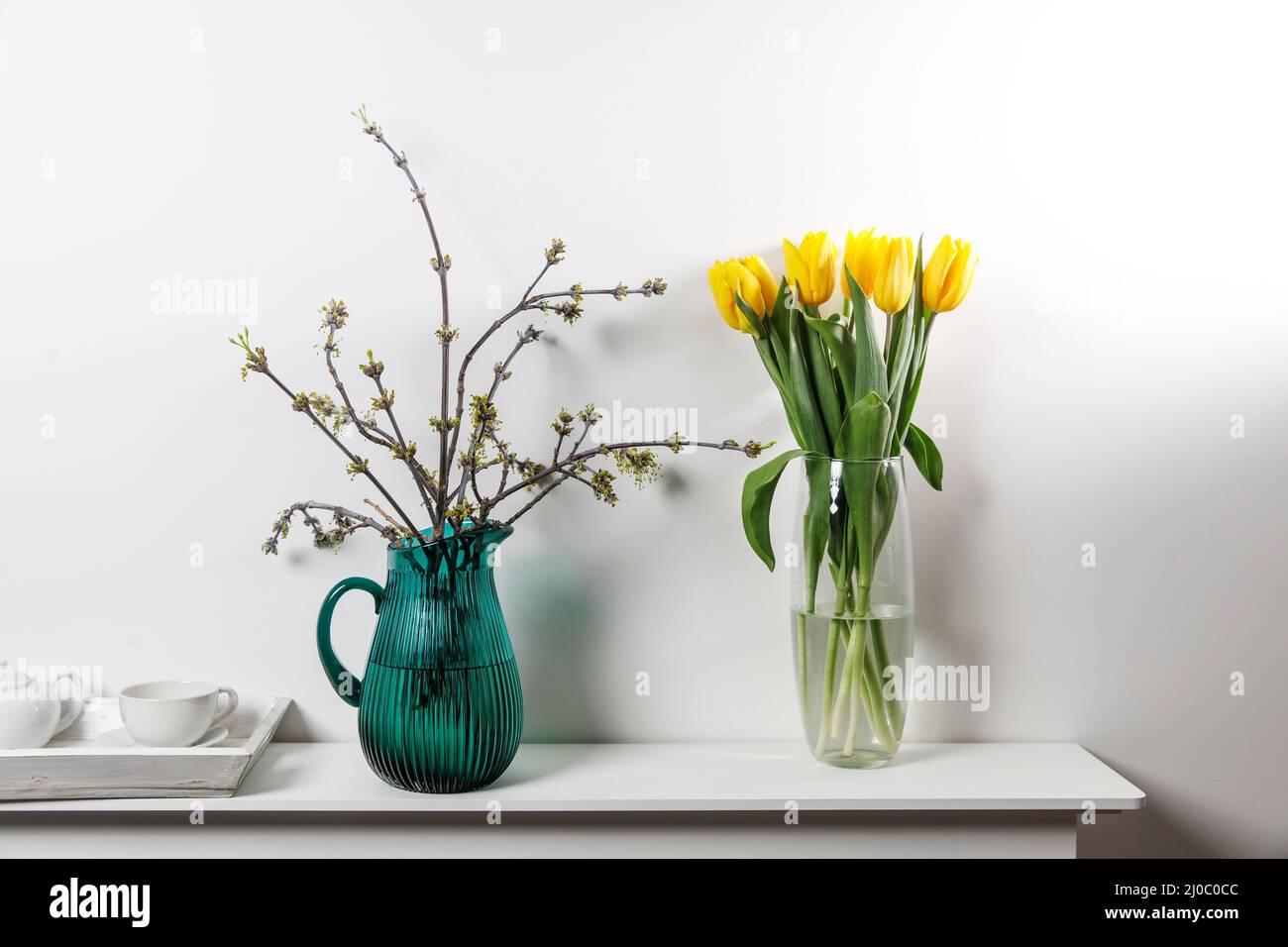 Bouquet of yellow tulip in the glass vase and American maple branches in green glass jar on white table Stock Photo