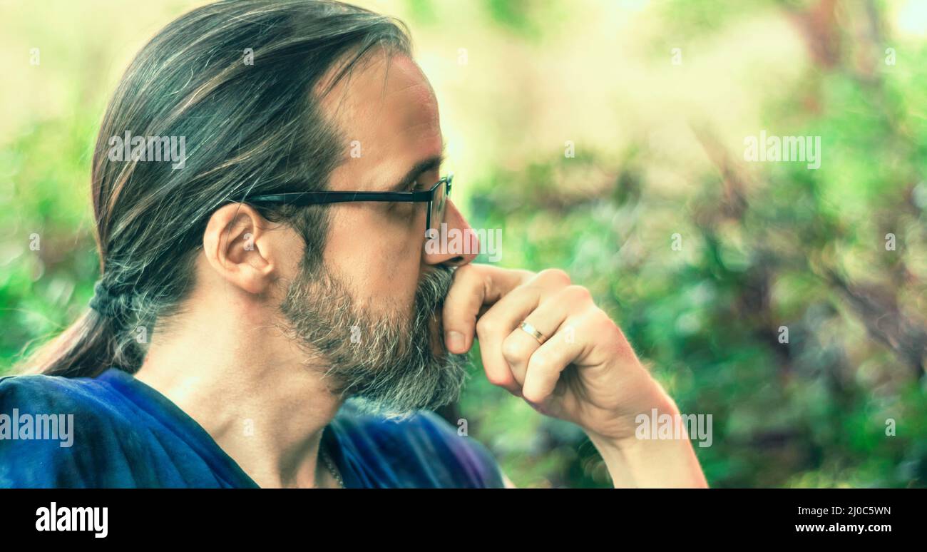 Portrait of one mid adult man with beard, glasses and long hair in a ponytail, looking thoughtful. Side view. Selective focus Stock Photo