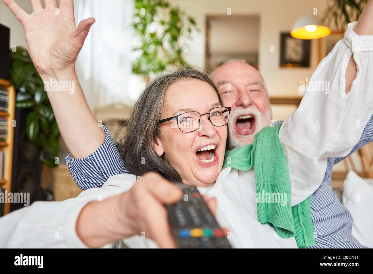 Couple of seniors with tv remote control cheering happily symbolizing lottery win or good luck Stock Photo