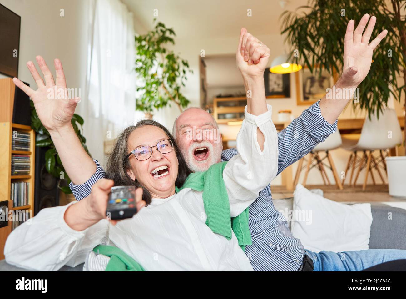 Cheering senior couple using remote control on sofa at home as lucky lottery winners Stock Photo