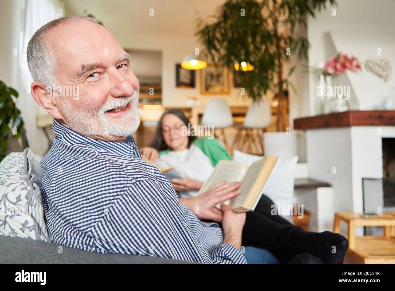 Happy retired senior man reading book in living room with wife on background Stock Photo