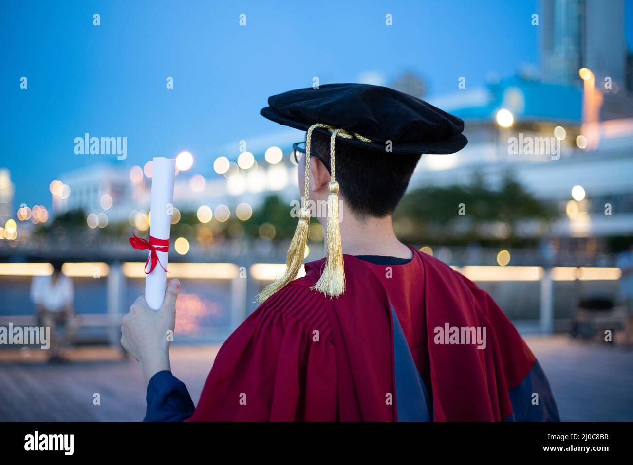 happy proud PhD graduated male students in Academic dress gown with certificate facing colorful city nights. back view Stock Photo