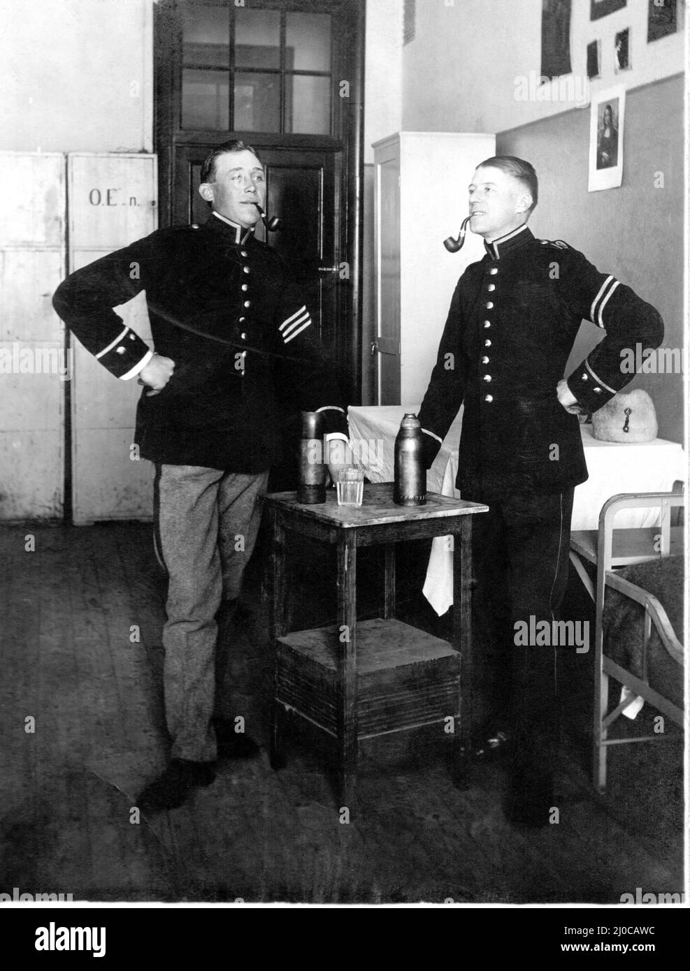 Authentic vintage photograph of two soldiers hands on hips smoking pipes, standing next to drinking flasks on table, Sweden. Concept of togetherness Stock Photo