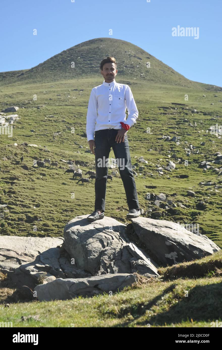 A handsome Indian young guy with wearing white shirt and black jean, looking at camera while standing over rock in the mountain. Stock Photo