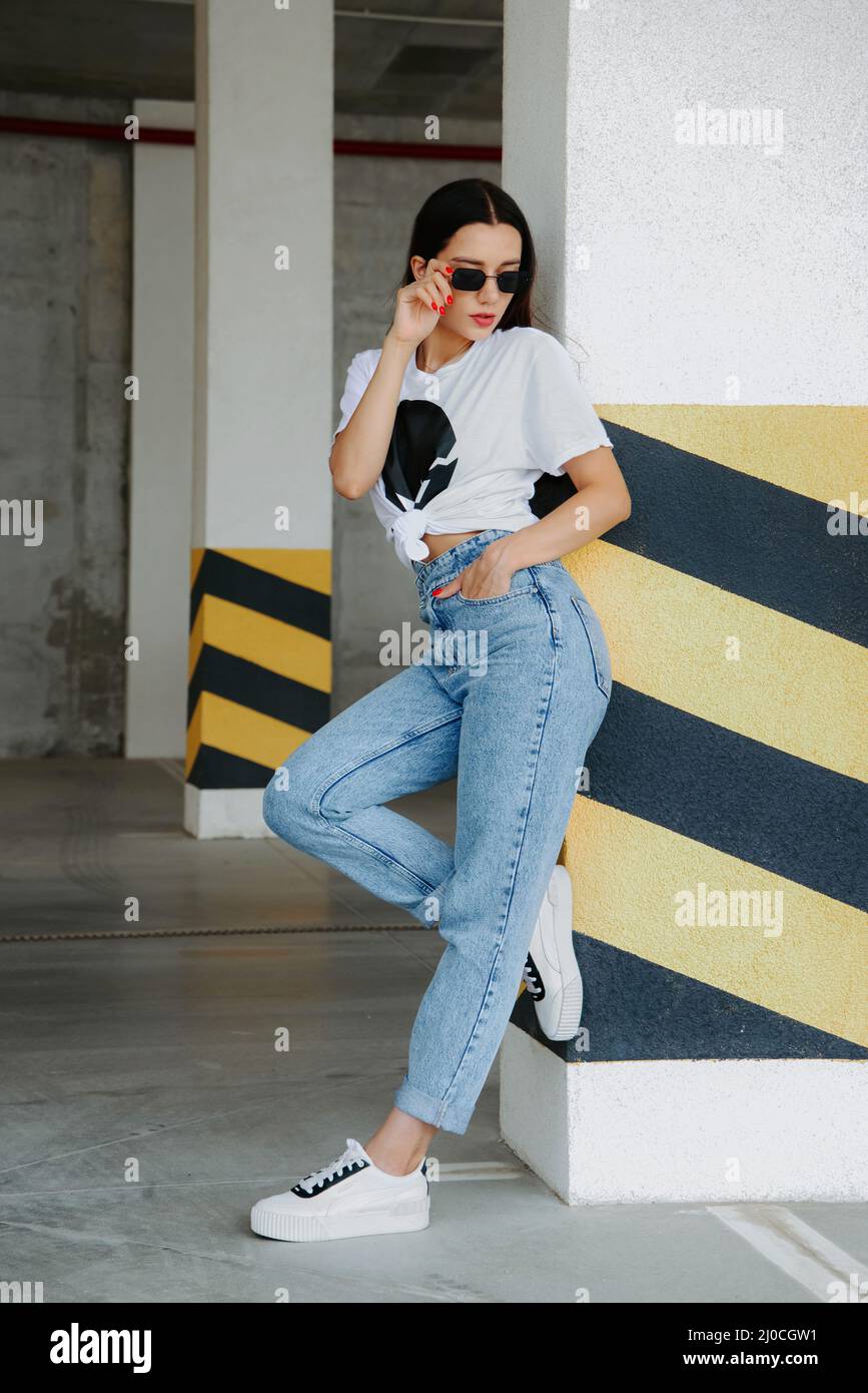 Pretty brunette girl wearing a t-shirt, jeans, sneakers and sunglasses,  stands in a car parking, near yellow-black stripes on a pillar Stock Photo  - Alamy