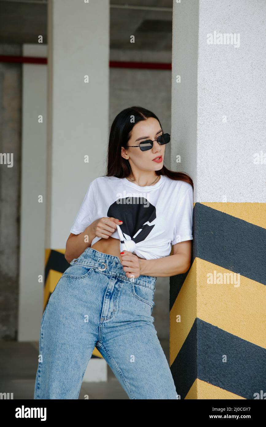 Pretty brunette girl wearing a t-shirt, jeans, sneakers and sunglasses, stands in a car parking, near yellow-black stripes on a pillar Stock Photo