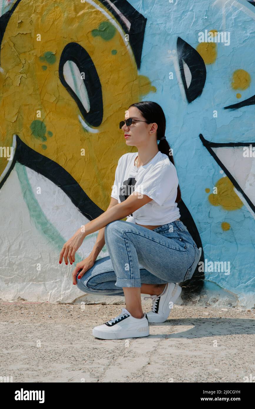 Pretty brunette girl wearing a t-shirt, jeans, sneakers and sunglasses, squats in front of graffiti on a wall on a sunny day Stock Photo