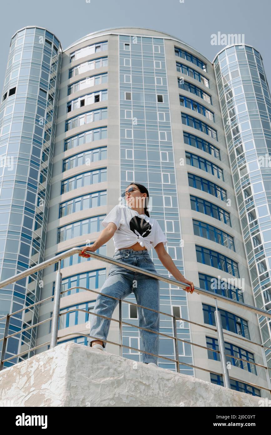 Pretty brunette girl, wearing a t-shirt and jeans, stands in front of a modern, shiny highrise, holding a railing; it is sunny & she wears sunglasses Stock Photo
