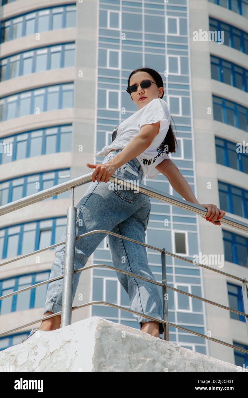 Pretty brunette girl, wearing a t-shirt and jeans, stands against railing in front of a modern, shiny highrise; it is sunny and she wears sunglasses Stock Photo