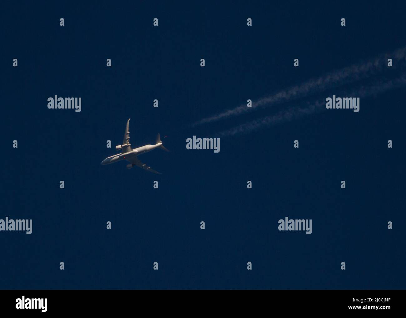 A commercial airliner with contrails at high altitude flies through a deep blue sky at sunset. Stock Photo