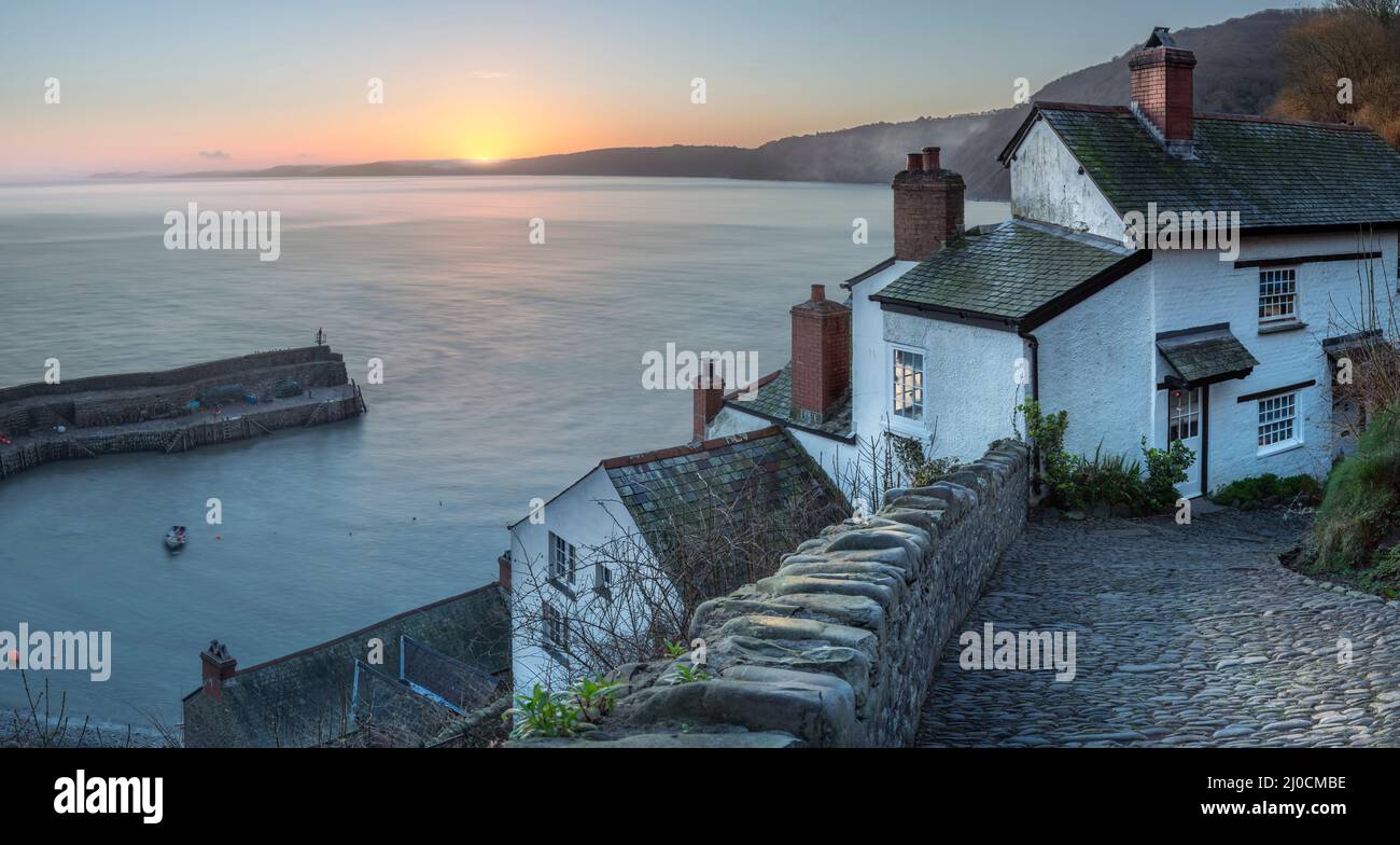 Mist rolls over the distant hills as the sun appears above the horizon, lighting up the whitewashed cottages that line the cobbled high street as it w Stock Photo