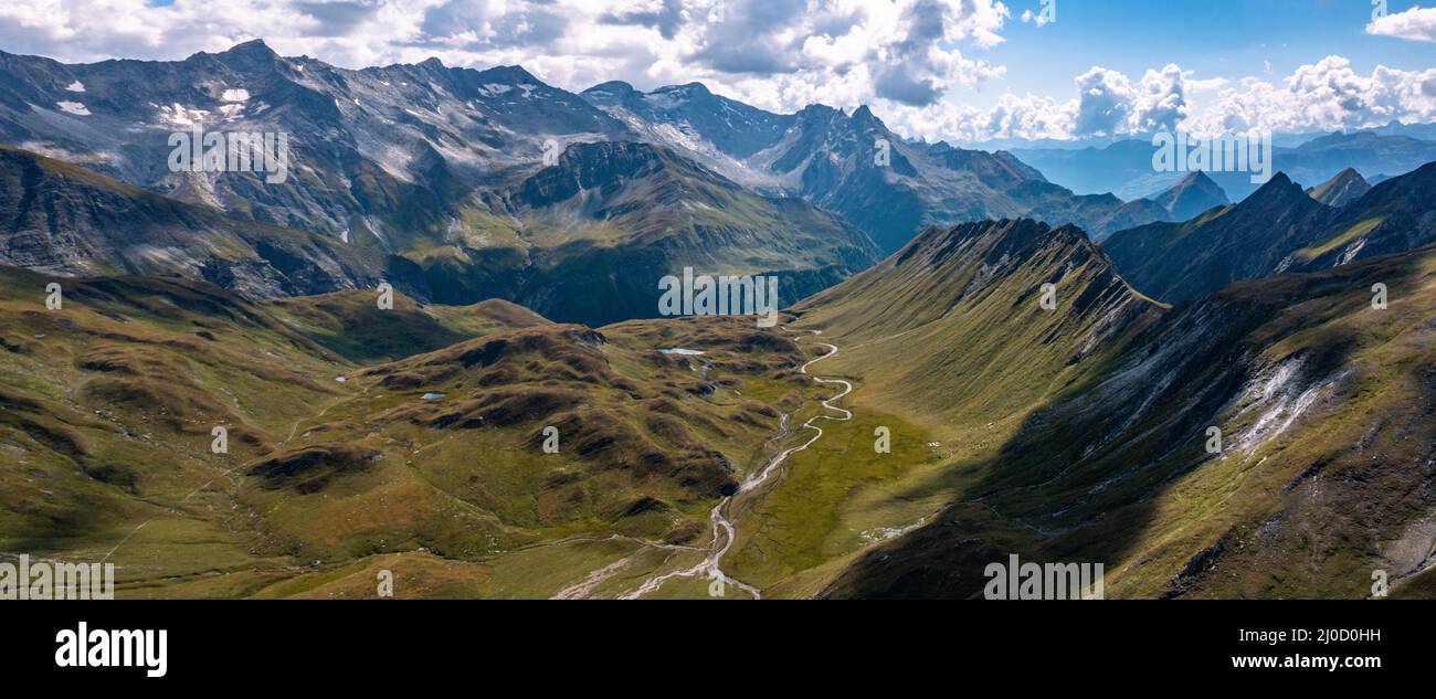 Aerial pano view towards Capanna Motterascio, an alpine hut on the Greina plateau in Blenio, Swiss Alps. A rocky ridge on the right leads the eye towards the river that flows sinuously in the valley. Stock Photo