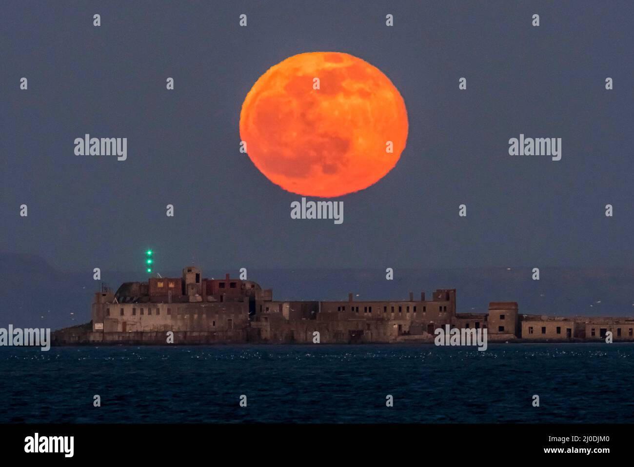 Weymouth, Dorset, UK.  18th March 2022.  UK Weather.  The full Worm Moon glows red as it rises up from behind the historic 19th century Portland Breakwater Fort on the outer breakwater of Portland Harbour near Weymouth in Dorset.  The now derelict fort also known as Chequered Fort was constructed between 1868 and 1878 and is Grade II listed.  Picture Credit: Graham Hunt/Alamy Live News Stock Photo