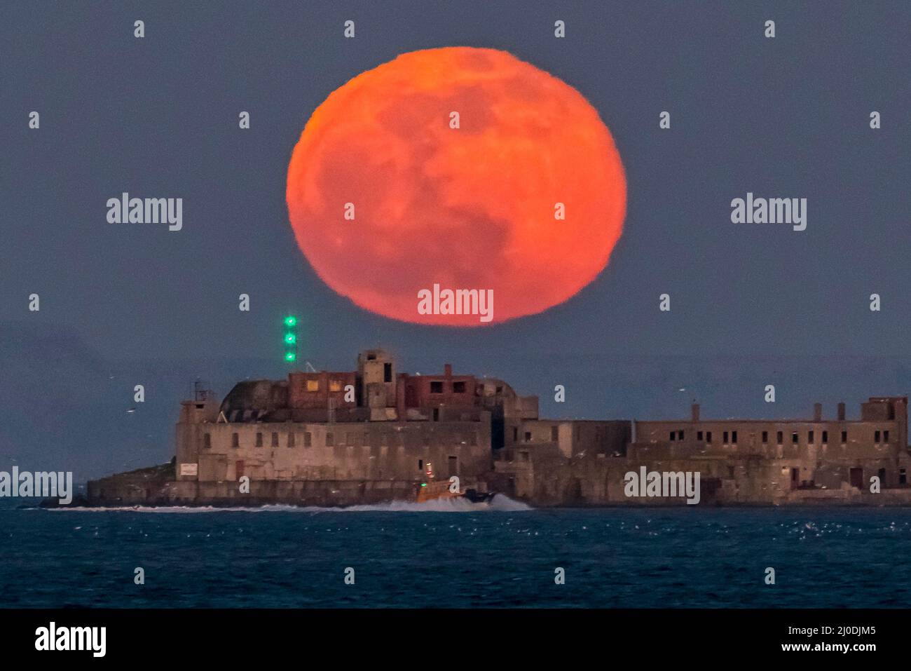 Weymouth, Dorset, UK.  18th March 2022.  UK Weather.  The full Worm Moon glows red as it rises up from behind the historic 19th century Portland Breakwater Fort on the outer breakwater of Portland Harbour near Weymouth in Dorset.  The now derelict fort also known as Chequered Fort was constructed between 1868 and 1878 and is Grade II listed.  Picture Credit: Graham Hunt/Alamy Live News Stock Photo