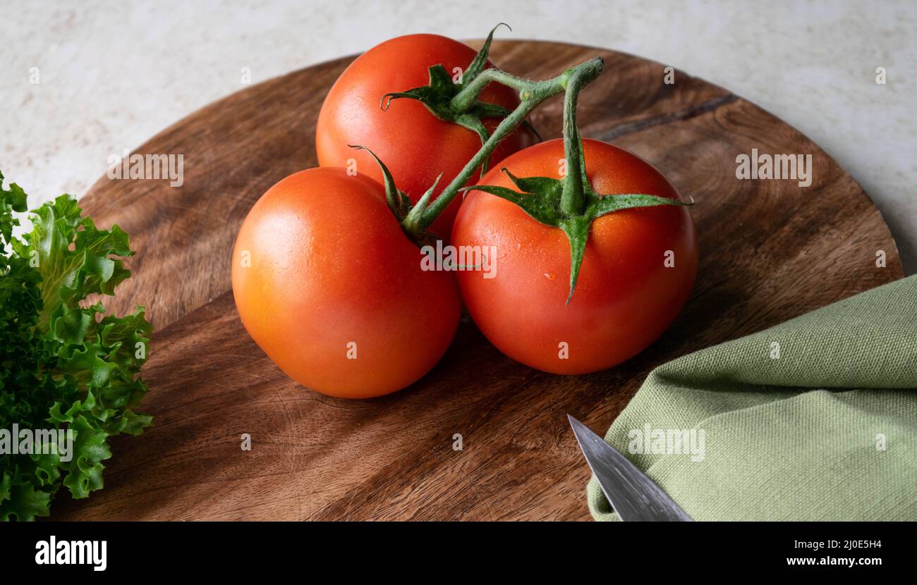 3 red tomatoes and a bunch of green lettuce on a round wooden cutting ...